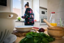 Анонимная женщина в разноцветной куртке стирает свежие овощи под чистой водой над раковиной на кухне дома — стоковое фото