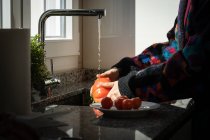 Обрізане зображення жінки в барвистій куртці, що миє свіжий помідор під чистою водою над раковиною на кухні вдома — стокове фото