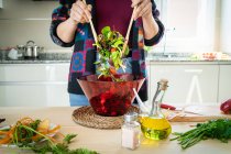 Imagem cortada de mulher em casaco multicolorido misturando legumes na tigela enquanto cozinha salada saudável na cozinha — Fotografia de Stock
