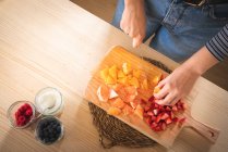 Imagen recortada de la mujer en traje casual picando frutas para una bebida saludable de naranja y fresa en la cocina en casa - foto de stock