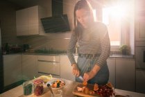 Junge Frau in lässigem Outfit schneidet frisches Obst, während sie in der gemütlichen Küche unter Sonnenstrahlen kocht — Stockfoto