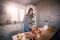 Jeune femme en tenue décontractée hacher des fruits frais tout en cuisinant dans une cuisine confortable sous les rayons de soleil lumineux — Photo de stock