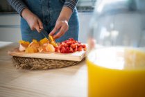 Imagem cortada de mulher em roupa casual cortar frutas para laranja saudável e bebida de morango na cozinha em casa — Fotografia de Stock