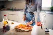 Immagine ritagliata di donna in abito casual tagliare frutta per una sana bevanda di arancia e fragola in cucina a casa — Foto stock