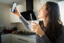 Молода жінка з банкою здорового йогурту посміхається і позує для селфі на кухні — стокове фото
