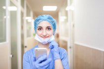 Cirurgiã feminina de pé no corredor enquanto verifica mensagens em seu telefone inteligente — Fotografia de Stock