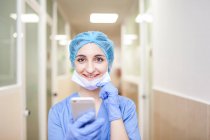 Жіночий хірург, що стоїть в коридорі під час перевірки повідомлень на смартфоні, подивіться на камеру і посміхніться — стокове фото