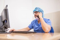 Cirujano masculino usando su computadora y su teléfono inteligente en su oficina antes de la cirugía - foto de stock