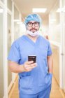 Männlicher Chirurg steht auf dem Flur, während er Nachrichten auf seinem Smartphone checkt, schaut in die Kamera — Stockfoto