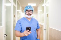 Мужчина-хирург стоит в коридоре, проверяя сообщения на смартфоне, смотрит в камеру — стоковое фото