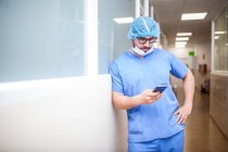 Male surgeon leaning against corridor wall while checking messages on his smart phone — Stock Photo