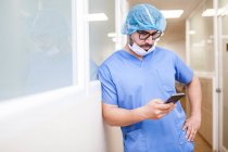 Чоловік хірург спирається на коридорну стіну під час перевірки повідомлень на смартфоні — стокове фото