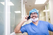 Мужской хирург опирается на стену коридора, разговаривая со своим смартфоном — стоковое фото