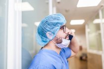 Chirurgo maschio appoggiato sulla parete del corridoio mentre parla con il suo smartphone, vista laterale — Foto stock