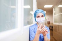 Жіночий хірург, що стоїть в коридорі під час перевірки повідомлень на смартфоні, подивіться на камеру і посміхніться — стокове фото