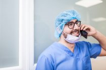 Cirujano masculino apoyado en la pared del pasillo mientras habla con su teléfono inteligente - foto de stock