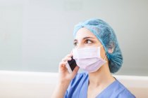 Female surgeon leaning on corridor wall while talking with her smart phone — Stock Photo