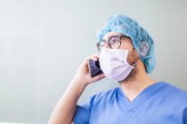 Männlicher Chirurg lehnt an Flurwand, während er mit seinem Smartphone spricht — Stockfoto