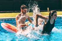 Jovem mulher salpicando no homem sentado no leito de ar na piscina — Fotografia de Stock
