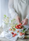 Сливочный сладкий десерт со свежей сочной клубникой, подаваемый в стакане на деревенском деревянном столе с печеньем и женской рукой, берущей ягоды на заднем плане — стоковое фото