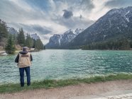 Вид на взрослого путешественника, фотографирующего горный хребет, стоя на берегу спокойного озера на природе — стоковое фото