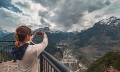 Back view woman in warm clothes standing on pier and looking at mountain ridge and taking picture with mobile phone in countryside — Stock Photo