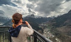 Rückansicht Frau in warmer Kleidung steht auf Pier und schaut auf Bergrücken und fotografiert mit Handy in der Landschaft — Stockfoto