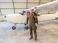 Selbstbewusster Pilot steht neben Retro-Flugzeug im Hangar und hält Helm — Stockfoto