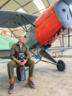 Серьезный мужчина-пилот в форме сидит рядом со старинным красочным бипланом в легком ангаре — стоковое фото