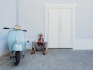 Hipster de meia-idade positivo em roupas elegantes perto de moto retro sentado no chão e usando smartphone em um dia ensolarado — Fotografia de Stock