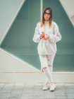Jovem hipster feminino moderno sonhador em roupas brancas casuais segurando fones de ouvido amarelo brilhante no fundo da parede — Fotografia de Stock