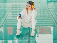 Jovem moderno hipster feminino em roupas casuais no fundo de óculos geométricos ouvindo música com smartphone e olhando para longe — Fotografia de Stock