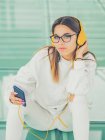 Young modern female hipster in casual clothes on background of geometric glasses listening music with smartphone and looking at camera — Stock Photo