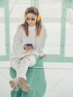 Joven hipster femenino moderno en ropa casual sobre fondo de gafas geométricas escuchando música con smartphone - foto de stock