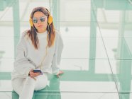 Молода сучасна жіноча хіпстерка в повсякденному одязі на фоні геометричних окулярів, слухаючи музику зі смартфоном — стокове фото
