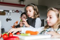 Маленькие девочки и мальчик режут и чистят спелые овощи, готовя здоровый салат на кухне вместе — стоковое фото
