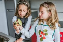 Маленька дівчинка демонструє половину стиглого авокадо своїй сестрі, стоячи на кухні, дивлячись на камеру — стокове фото