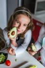 Kleines Mädchen demonstriert der Kamera die Hälfte der reifen Avocado, während es in der Küche steht und in die Kamera schaut — Stockfoto