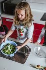 Kleines Mädchen wäscht frische Kräuter im Sieb unter klarem Wasser, während es in der Küche Salat zubereitet — Stockfoto