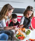 Маленькі дівчата і хлопчик ріжуть і очищають стиглі овочі, готуючи здоровий салат на кухні разом — стокове фото