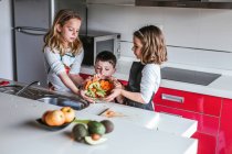 Маленькие девочки и мальчик режут и чистят спелые овощи, готовя здоровый салат на кухне вместе — стоковое фото