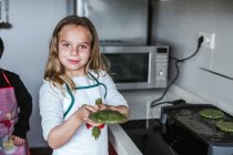 Kleines Mädchen lächelt in die Kamera und hält Teller mit grünen vegetarischen Schnitzeln in der heimischen Küche — Stockfoto