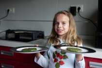 Niña sonriendo y mirando a la cámara mientras sostiene platos con chuletas vegetarianas verdes en la cocina en casa - foto de stock