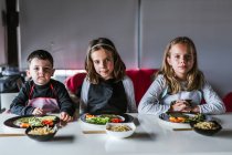 Niño y dos niñas esperando para comer sabrosos fideos con chuletas vegetarianas y verduras mientras están sentados en la mesa en casa - foto de stock