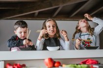 Мальчик и две девочки едят вкусную лапшу с вегетарианскими котлетами и овощами, сидя за столом дома — стоковое фото