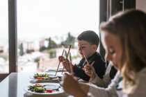 Хлопчик і дві дівчини насолоджуються смачною локшиною з вегетаріанськими котлети та овочі, сидячи за столом вдома — стокове фото