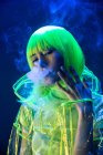 Young pretty unusual Asian woman in plastic transparent raincoat and yellow hair smoking in fluorescent light — Stock Photo