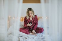 Молодая красивая женщина в пижаме сидит на кровати и использует смартфон — стоковое фото