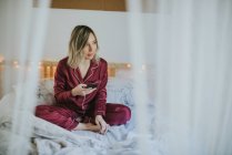 Jeune jolie femme en pyjama assise sur le lit et utilisant un smartphone — Photo de stock