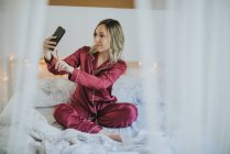 Молода красива жінка в піжамі сидить на ліжку, приймаючи селфі зі смартфоном — стокове фото